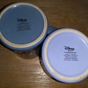 May include: Two ceramic Disney Store mugs with light blue and lavender bottoms. The mugs have a white rim and the Disney Store logo is printed on the bottom. The text "Made in Thailand" is also printed on the bottom of the mugs.