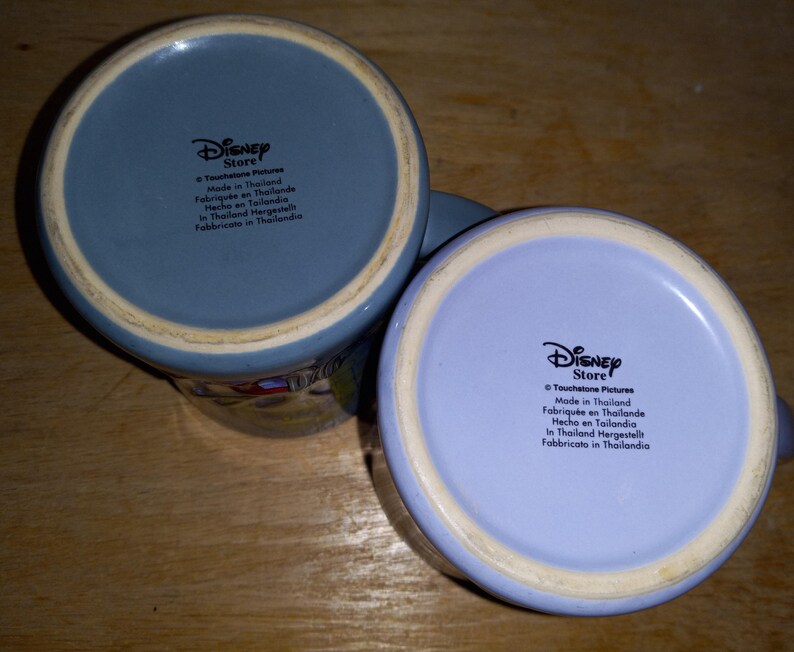 May include: Two ceramic Disney Store mugs with light blue and lavender bottoms. The mugs have a white rim and the Disney Store logo is printed on the bottom. The text "Made in Thailand" is also printed on the bottom of the mugs.