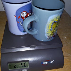 May include: Two ceramic mugs with character designs, one light blue and one gray, sit on a digital scale. The scale displays a weight of 2.5 ounces. The mugs feature images of characters from a popular animated film.