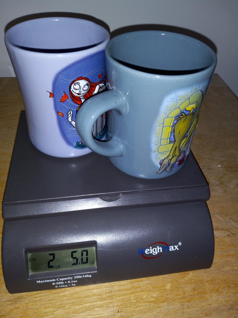 May include: Two ceramic mugs with character designs, one light blue and one gray, sit on a digital scale. The scale displays a weight of 2.5 ounces. The mugs feature images of characters from a popular animated film.