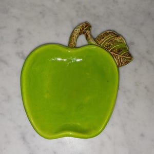 May include: A bright lime green ceramic dish shaped like an apple. The dish features a glossy finish and a textured, brown-toned stem and leaf attached to the top right side, providing a realistic detail to the overall apple-shaped design.