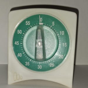 May include: A square white kitchen timer with a circular green face featuring white numbers from 0 to 55 in five-minute increments. A central translucent gray knob sits in the middle of the dial, which is encased in a clear plastic cover.