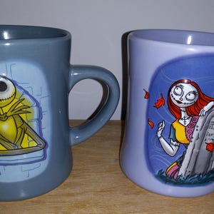 May include: Two ceramic mugs featuring characters from The Nightmare Before Christmas. One mug is gray with Jack Skellington, the other is lavender with Sally. Both mugs have curved handles and character illustrations.