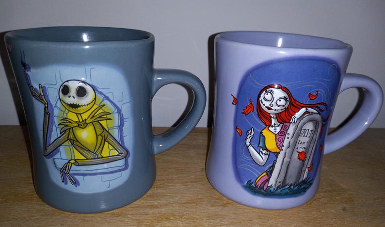 May include: Two ceramic mugs featuring characters from The Nightmare Before Christmas. One mug is gray with Jack Skellington, the other is lavender with Sally. Both mugs have curved handles and character illustrations.