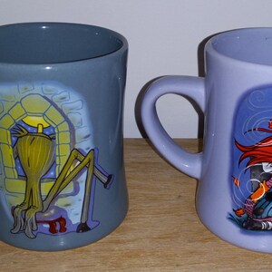 May include: Two ceramic mugs with curved handles. One is gray with a yellow and black cartoon character in a window. The other is lavender with a red-haired cartoon character. Both mugs have a glossy finish.