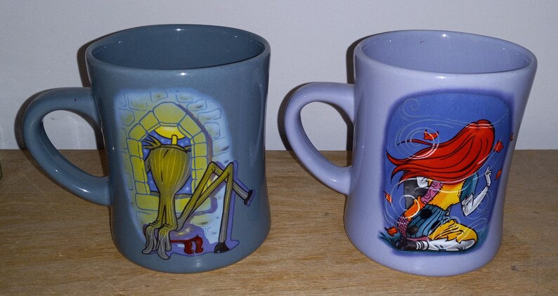 May include: Two ceramic mugs with curved handles. One is gray with a yellow and black cartoon character in a window. The other is lavender with a red-haired cartoon character. Both mugs have a glossy finish.