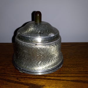 May include: A silver-toned, bell-shaped container with a lid and a small, dark brown knob. The container has a textured surface and sits on a wooden surface. The lid is slightly domed, and the knob is cylindrical.