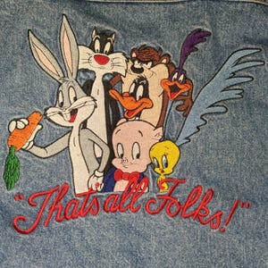 May include: A blue denim jacket featuring an embroidered graphic of Looney Tunes characters including Bugs Bunny holding a carrot, Sylvester, Daffy Duck, Taz, Road Runner, Porky Pig, and Tweety Bird, with the red embroidered text That's all Folks!
