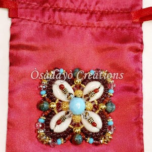 May include: A burgundy satin pouch with a decorative floral design. The design features white seashells, turquoise beads, and gold accents. The text "Osadayo Creations" is printed on the pouch.