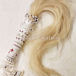 May include: A decorative white and red beaded ceremonial item with a long, flowing, light-colored hair tail. The handle is intricately adorned with beads and patterns. The text "Osadayo Creations" is visible on the item.