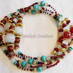 May include: A multi-strand necklace featuring a variety of beads in white, red, brown, turquoise, and gold. The necklace includes large, round white beads, smaller red and brown beads, and gold-colored accents. The text "Òsadayò Creations" is visible.