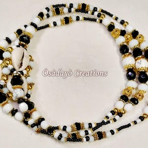 May include: A handcrafted necklace featuring a combination of white, black, and gold beads. The necklace incorporates cowrie shells and decorative gold accents. The text "Òsàdàyò Creations" is visible on the image.
