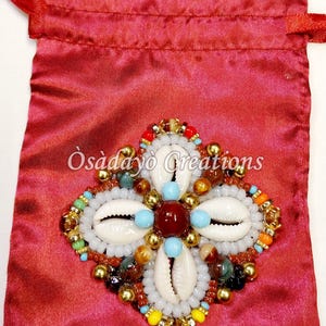 May include: A red satin drawstring bag with a beaded embellishment. The embellishment features white seashells, turquoise, red, and gold beads, and the text "Òsàdàyò Creations".