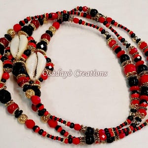 May include: A triple strand beaded necklace with red, black, and gold beads. The necklace features cowrie shells and is made by Osadayó Creations.