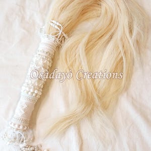 May include: A decorative white and cream-colored item with a long, flowing, light blonde hair-like section. The handle is wrapped in white fabric and adorned with beads and pearls. The text "Osadayo Creations" is visible.