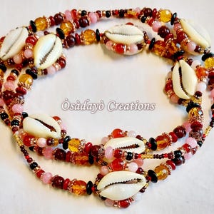 May include: A multi-strand necklace featuring cowrie shells and beads in shades of red, pink, amber, and black. The text "Òsàdàyò Creations" is visible.