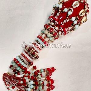 May include: A handcrafted, beaded ceremonial rattle. The red, bulbous head is adorned with white and gold accents. The handle and tassel feature red, white, and turquoise beads. The text "Osadayo Creations" is visible.