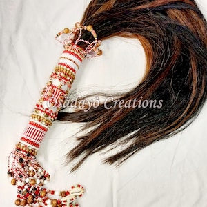 May include: A ceremonial beaded wand with a long, flowing hair tail. The wand features a white, red, and gold beaded handle, adorned with cowrie shells and decorative elements. The text "Osadayo Creations" is visible.