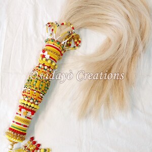 May include: A long, blonde hair tassel with a handle decorated with colorful beads and a tassel at the bottom. The handle is made of yellow, red, green, and white beads.