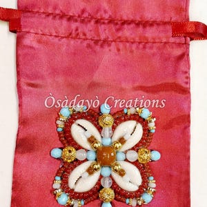 May include: A red satin drawstring bag with a beaded floral design. The design features white seashells, turquoise beads, and gold accents. The text "Òsàdàyò Creations" is printed on the bag.