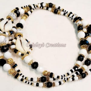 May include: A triple-strand necklace with white, black, and gold beads. The necklace features cowrie shells and is made with a variety of bead sizes and textures. The text "Osadayó Creations" is visible in the image.