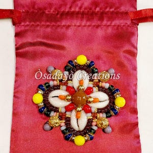 May include: A burgundy satin drawstring bag with a beaded embellishment. The embellishment features white seashells, yellow, red, and blue beads, and the text "Osadayo Creations".