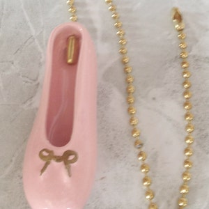 Könnte beinhalten: Ein rosa Keramik-Ballerinaschuh mit einer goldenen Schleife und einer daran befestigten goldenen Kette. Die Kette besteht aus kleinen, runden Gliedern.