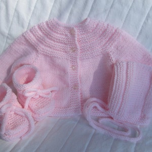 May include: A pink knitted baby outfit set including a cardigan, hat, and booties.