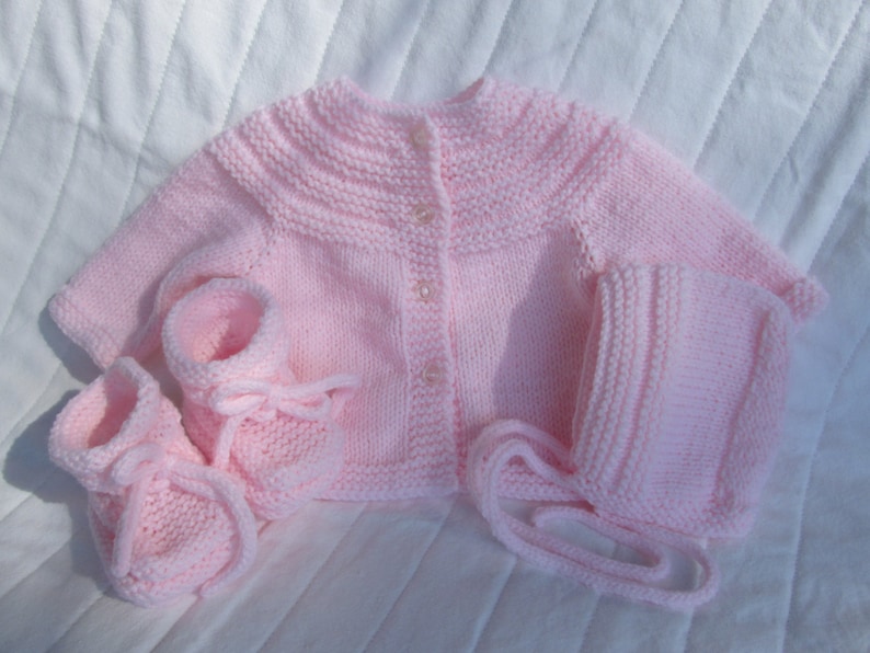 May include: A pink knitted baby outfit set including a cardigan, hat, and booties.