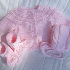 May include: A pink knitted baby cardigan, hat, and booties set. The cardigan has a button closure and the hat has a matching tie.