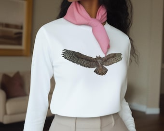 Può includere: Una maglia bianca a maniche lunghe con una dettagliata illustrazione di un gufo con le ali spiegate. La maglia è abbinata a pantaloni beige, una sciarpa rosa e un berretto da baseball beige. Orecchini a cerchio dorati completano il look.