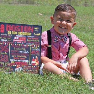 K&ouml;nnte beinhalten: Ein Junge in einem rosafarbenen Hemd, Fliege und Hosentr&auml;ger sitzt auf einer gr&uuml;nen Wiese. Er h&auml;lt eine schwarze Tafel mit dem Text "BOSTON My 3rd Birthday" und Details zu seinen Lieblingsspielzeugen, Dingen, die er liebt, sowie seinem Alter und Gewicht.