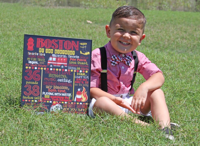 K&ouml;nnte beinhalten: Ein Junge in einem rosafarbenen Hemd, Fliege und Hosentr&auml;ger sitzt auf einer gr&uuml;nen Wiese. Er h&auml;lt eine schwarze Tafel mit dem Text "BOSTON My 3rd Birthday" und Details zu seinen Lieblingsspielzeugen, Dingen, die er liebt, sowie seinem Alter und Gewicht.