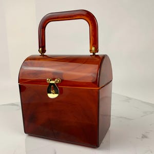 May include: A vintage-style, brown acrylic handbag with a curved handle and a gold-colored clasp. The boxy purse has a tortoiseshell pattern and a rectangular shape. The handle is attached with gold-colored hardware.