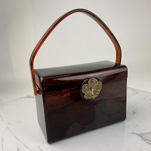 May include: A vintage rectangular handbag with a glossy, dark brown finish. It features a curved, amber-colored handle and a decorative gold-toned floral clasp. The bag's design is reminiscent of mid-century fashion.