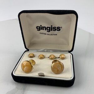 Puede incluir: Un juego de gemelos y botones de camisa dorados en una caja de terciopelo negro. Los gemelos y los botones tienen un patrón de remolino en color marrón claro. La caja tiene el texto "gingiss® CUSTOM COLLECTION" en la tapa interior.