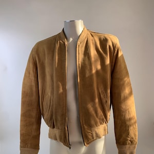 May include: A tan suede bomber jacket with a ribbed collar, cuffs, and hem. The jacket has a zipper closure and two front pockets. The jacket is displayed on a mannequin against a white background.