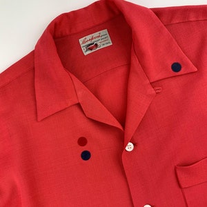 May include: A red short-sleeved button-down shirt with a white label that reads "Goodford Sport Shirt" and a small illustration of a car. The shirt has three buttons and a pocket on the left side. There are three small decorative buttons on the front of the shirt, one red and two blue.