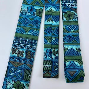 May include: A vintage necktie with a vibrant geometric and floral pattern in shades of blue, green, and brown. The tie features repeating shapes and stylized flower motifs, creating a retro aesthetic. The tie is displayed against a white background.