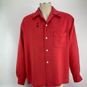 May include: A long-sleeved, red button-down shirt with a single pocket on the left chest. The shirt has a collar with two small, dark blue buttons.