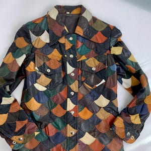May include: A long-sleeve leather jacket featuring a multi-colored scale pattern in shades of brown, green, blue, yellow, and cream. The jacket includes a pointed collar, a front button closure with silver-tone snaps, and two chest pockets.