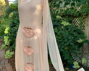 May include: A light peach-colored dress with a V-neck and a flowing skirt. The dress features a decorative belt and three floral embellishments. One long, sheer sleeve drapes down. The dress is displayed on a mannequin.
