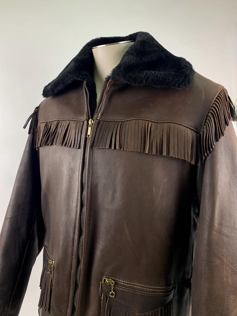 May include: Brown leather jacket with black faux fur collar and fringe detailing. The jacket features a zip-up front, two front pockets with fringe, and gold-tone hardware. A classic western-inspired outerwear piece.