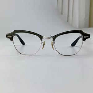 May include: Eyeglasses with a dark green upper frame and silver lower frame. The glasses have a classic, semi-rimless design with clear lenses and black temples. The bridge and nose pads are silver and gold.