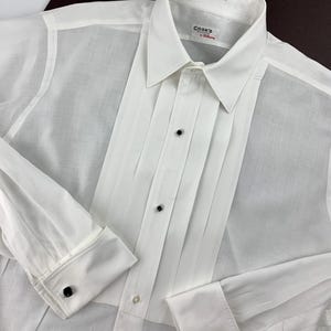 Pode incluir: Uma camisa branca com frente pregueada e punhos franceses. A camisa tem uma gola clássica e botões pretos. A etiqueta diz "Cook's by Hollywood". A camisa é feita de um tecido leve.