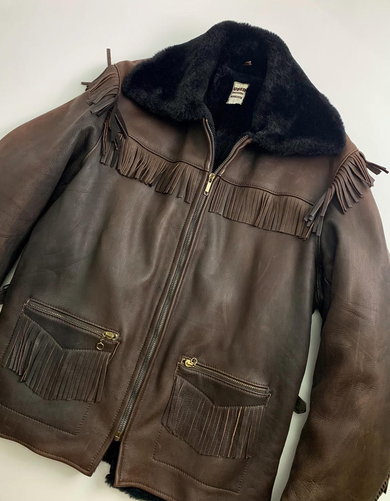 May include: A brown leather jacket with a black faux fur collar and fringe detailing. The jacket features a zipper closure, two front pockets with fringe, and a label on the inside. The jacket is a medium brown color.