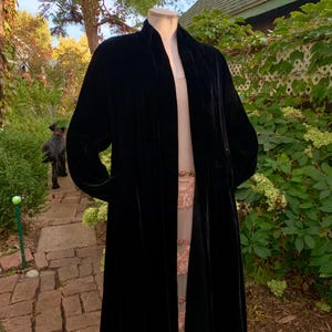 Op de afbeelding: Een lange, zwarte fluwelen jas met een vloeiend silhouet. De jas wordt buiten op een mannequin getoond, wat de elegante drapeering laat zien. De jas heeft een kraagloos ontwerp en lange mouwen.