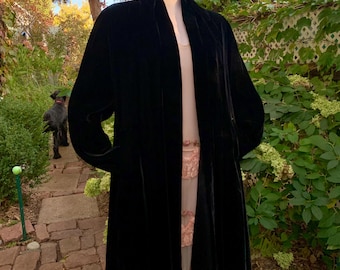May include: A long, black velvet coat with a flowing silhouette. The coat is displayed on a mannequin outdoors, showcasing its elegant drape. The coat has a collarless design and long sleeves, perfect for layering.