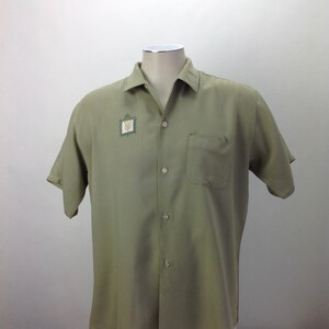 May include: A light green short-sleeved button-down shirt with a small embroidered design on the left chest. The shirt has a pocket on the left side.
