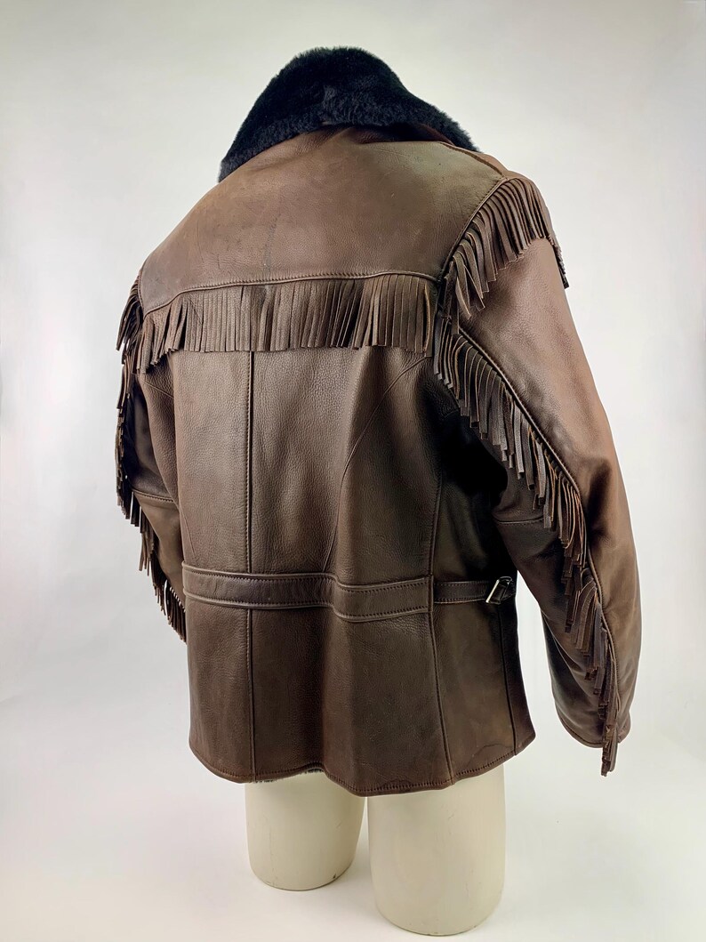 May include: Brown leather jacket with fringe detailing. The jacket features a black fur collar and a belt at the waist. The fringe is along the shoulders and sleeves. The jacket is displayed on a mannequin against a white background.